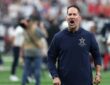 Cowboys head coach Brian Schottenheimer shouts in celebration as he walks off the field holding his cap.