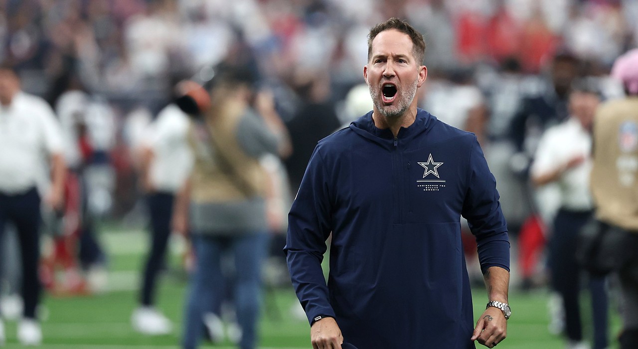 Cowboys head coach Brian Schottenheimer shouts in celebration as he walks off the field holding his cap.
