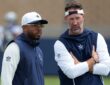 Dallas Cowboys defensive coordinator Christian Parker stands beside head coach Brian Schottenheimer, who talks with arms crossed.