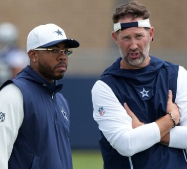 Dallas Cowboys defensive coordinator Christian Parker stands beside head coach Brian Schottenheimer, who talks with arms crossed.