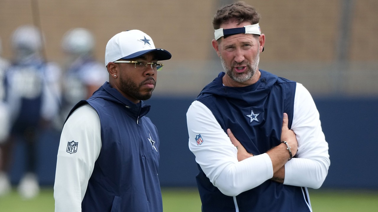 Dallas Cowboys defensive coordinator Christian Parker stands beside head coach Brian Schottenheimer, who talks with arms crossed.