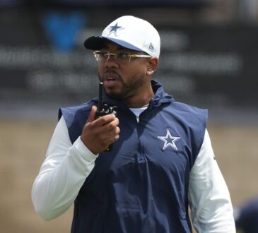 Dallas Cowboys defensive coordinator Christian Parker talks into a handheld radio while wearing a team cap and navy vest.