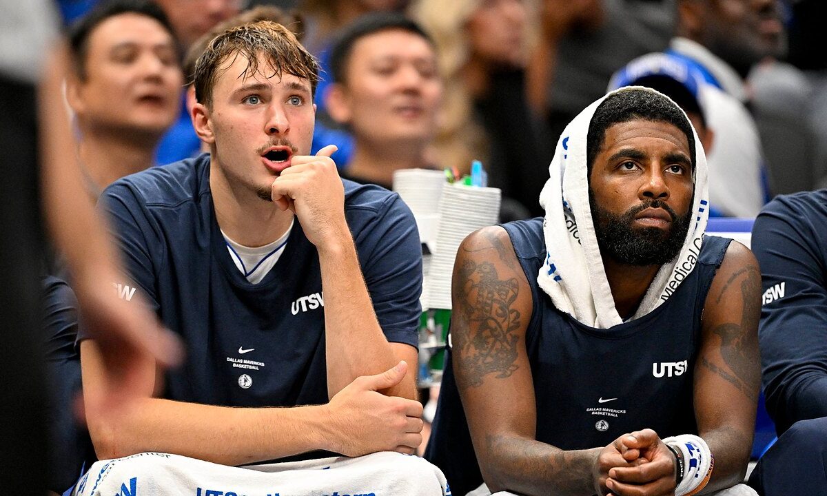 Dallas Mavericks forward Cooper Flagg reacts with his hand at his chin beside guard Kyrie Irving, who wears a towel over his head.