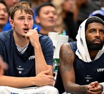 Dallas Mavericks forward Cooper Flagg reacts with his hand at his chin beside guard Kyrie Irving, who wears a towel over his head.