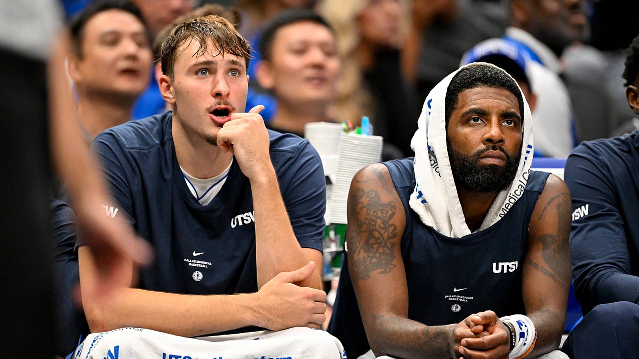 Dallas Mavericks forward Cooper Flagg reacts with his hand at his chin beside guard Kyrie Irving, who wears a towel over his head.