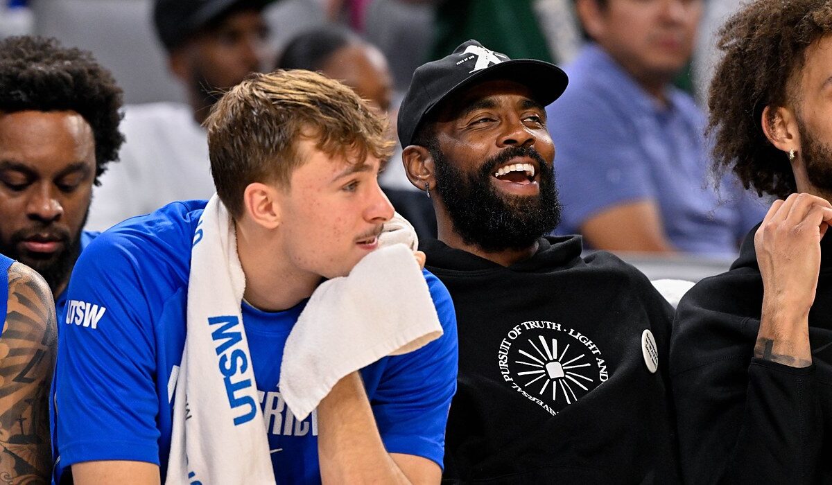 Mavericks forward Cooper Flagg rests his chin on a towel as guard Kyrie Irving laughs beside him on the bench.