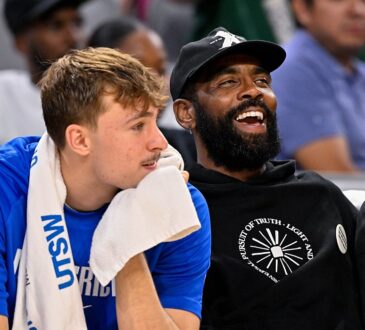 Mavericks forward Cooper Flagg rests his chin on a towel as guard Kyrie Irving laughs beside him on the bench.