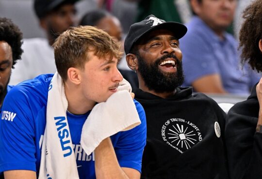 Mavericks forward Cooper Flagg rests his chin on a towel as guard Kyrie Irving laughs beside him on the bench.