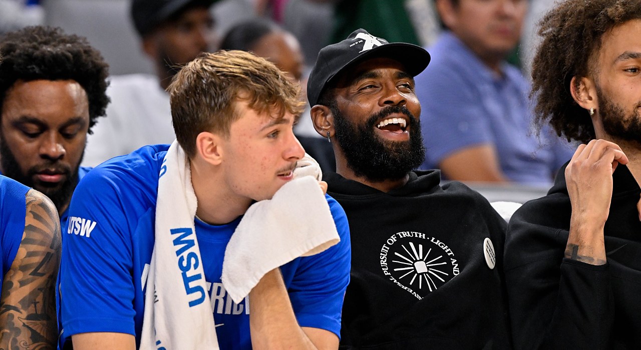 Mavericks forward Cooper Flagg rests his chin on a towel as guard Kyrie Irving laughs beside him on the bench.