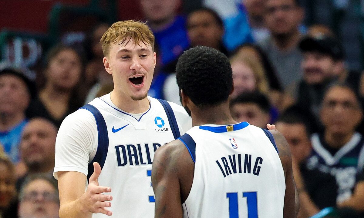 Dallas Mavericks forward Cooper Flagg smiles and reaches toward guard Kyrie Irving, who has his back to the camera.