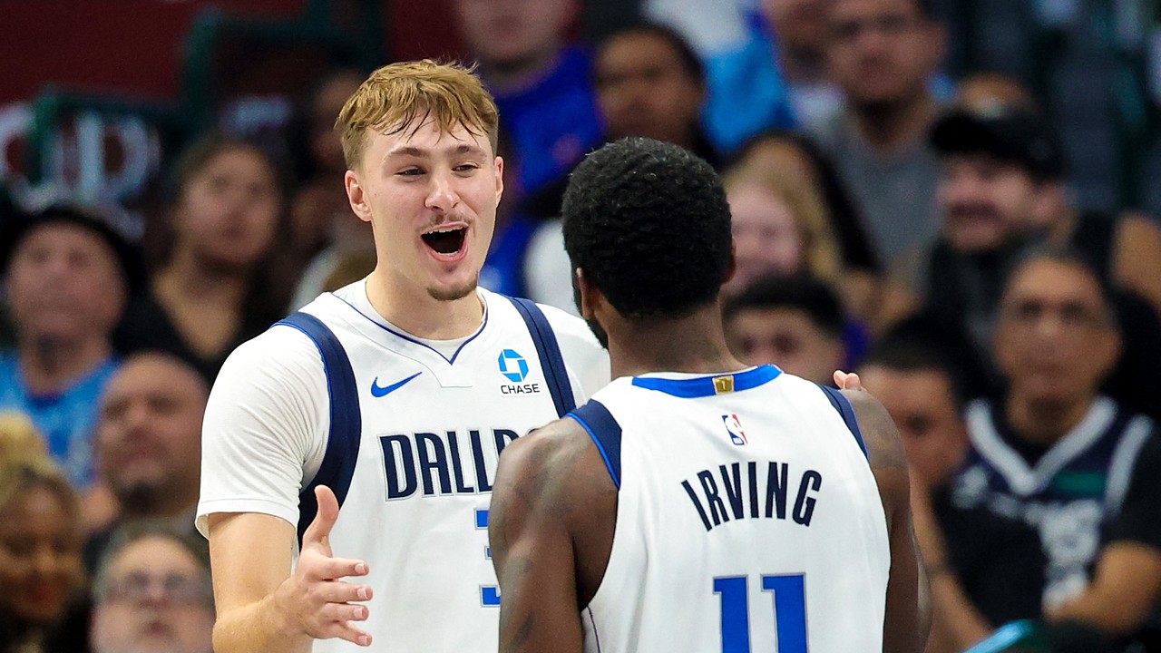 Dallas Mavericks forward Cooper Flagg smiles and reaches toward guard Kyrie Irving, who has his back to the camera.