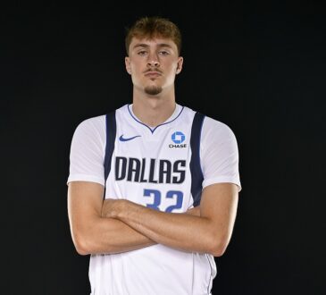 Dallas Mavericks forward Cooper Flagg stands with his arms crossed in a white Dallas jersey against a black backdrop.