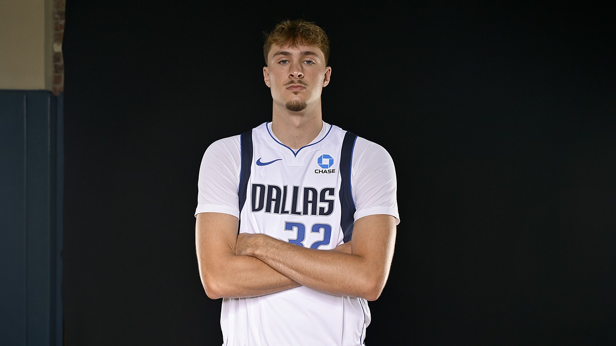 Dallas Mavericks forward Cooper Flagg stands with his arms crossed in a white Dallas jersey against a black backdrop.