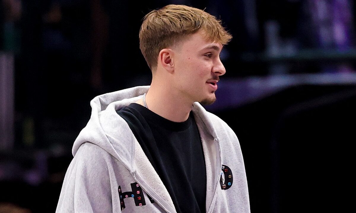 Dallas Mavericks forward Cooper Flagg stands courtside in a gray zip-up hoodie over a black T-shirt.