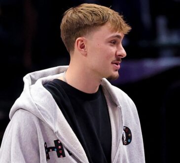 Dallas Mavericks forward Cooper Flagg stands courtside in a gray zip-up hoodie over a black T-shirt.