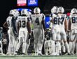 Dallas Cowboys players stand in a huddle with their backs to the camera, name bars and numbers visible.