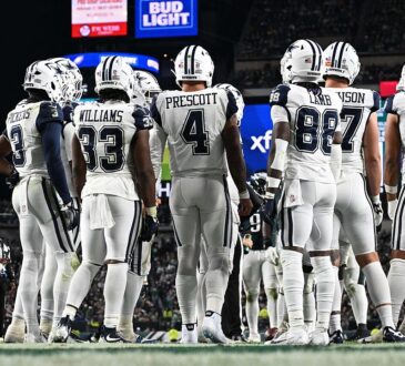 Dallas Cowboys players stand in a huddle with their backs to the camera, name bars and numbers visible.