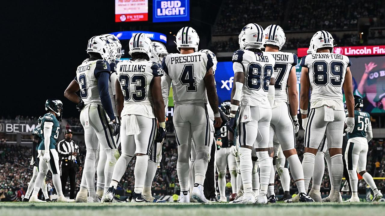 Dallas Cowboys players stand in a huddle with their backs to the camera, name bars and numbers visible.