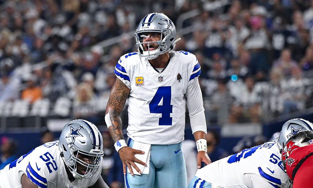 Dallas Cowboys quarterback Dak Prescott shouts at the line of scrimmage with a hand on his hip as linemen settle into stances.