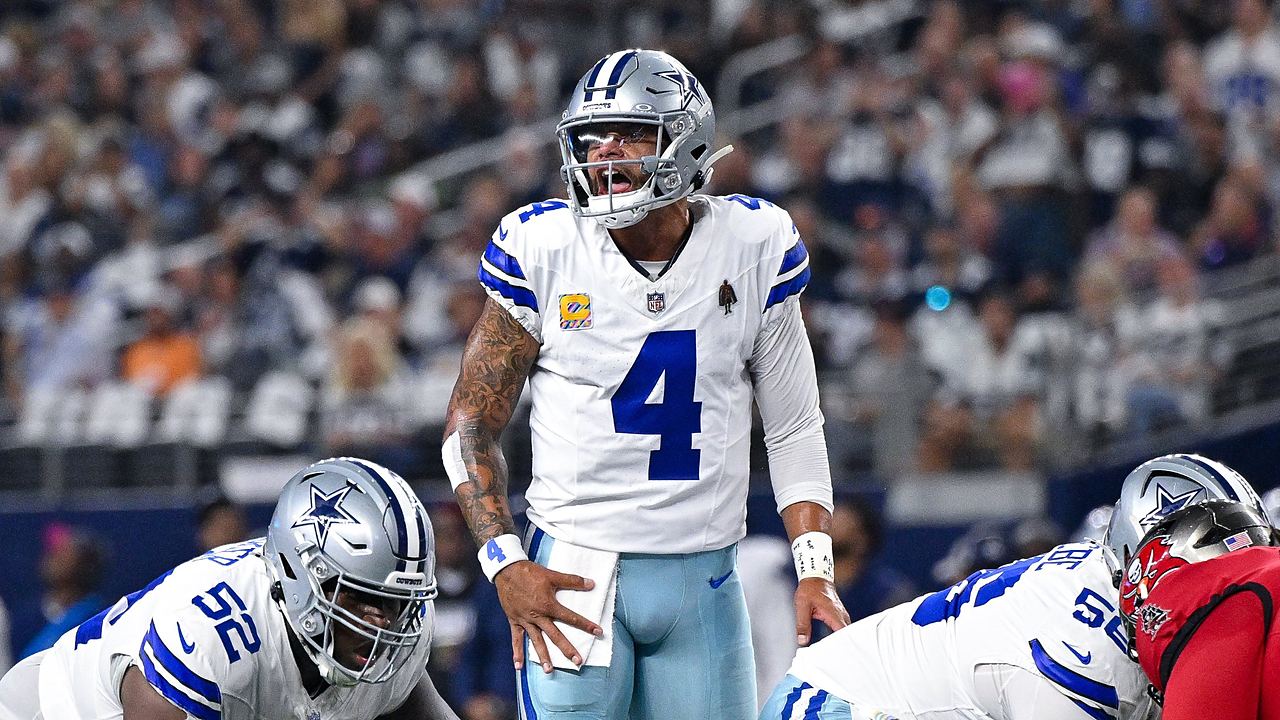 Dallas Cowboys quarterback Dak Prescott shouts at the line of scrimmage with a hand on his hip as linemen settle into stances.
