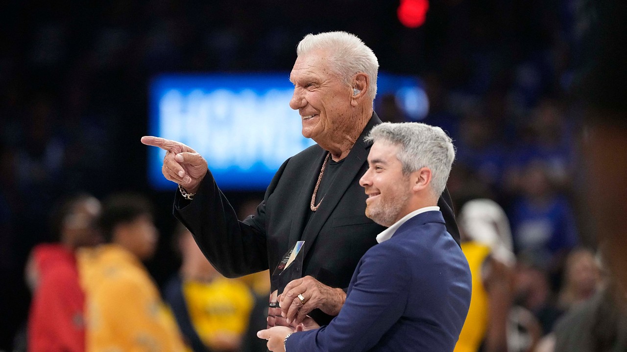 Former Dallas Mavericks coach Donald Nelson smiles and points while holding a glass award on the court.