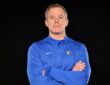 Mavericks head coach Dusty May poses with his arms crossed in a blue team quarter-zip against a black backdrop.