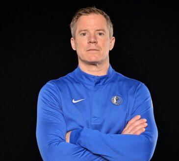 Mavericks head coach Dusty May poses with his arms crossed in a blue team quarter-zip against a black backdrop.