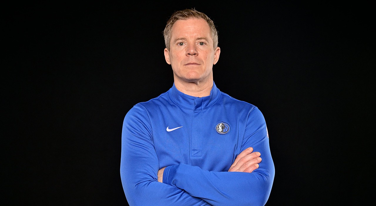 Mavericks head coach Dusty May poses with his arms crossed in a blue team quarter-zip against a black backdrop.