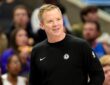 Dallas Mavericks head coach Dusty May smiles on the sideline in a black Mavericks sweater as fans watch behind him.