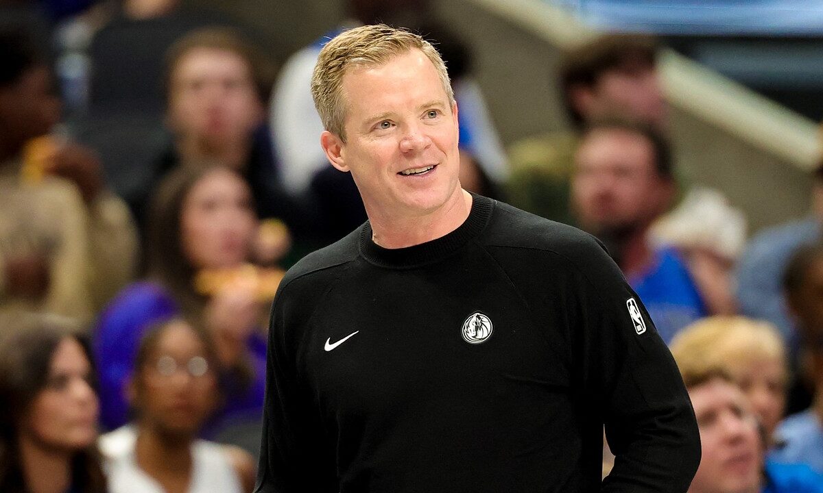Dallas Mavericks head coach Dusty May smiles on the sideline in a black Mavericks sweater as fans watch behind him.