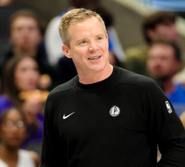 Dallas Mavericks head coach Dusty May smiles on the sideline in a black Mavericks sweater as fans watch behind him.