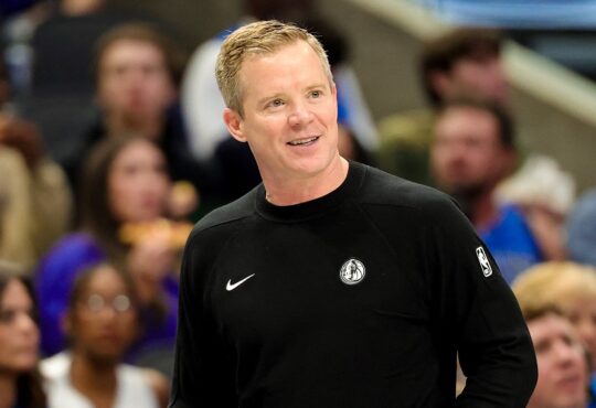 Dallas Mavericks head coach Dusty May smiles on the sideline in a black Mavericks sweater as fans watch behind him.