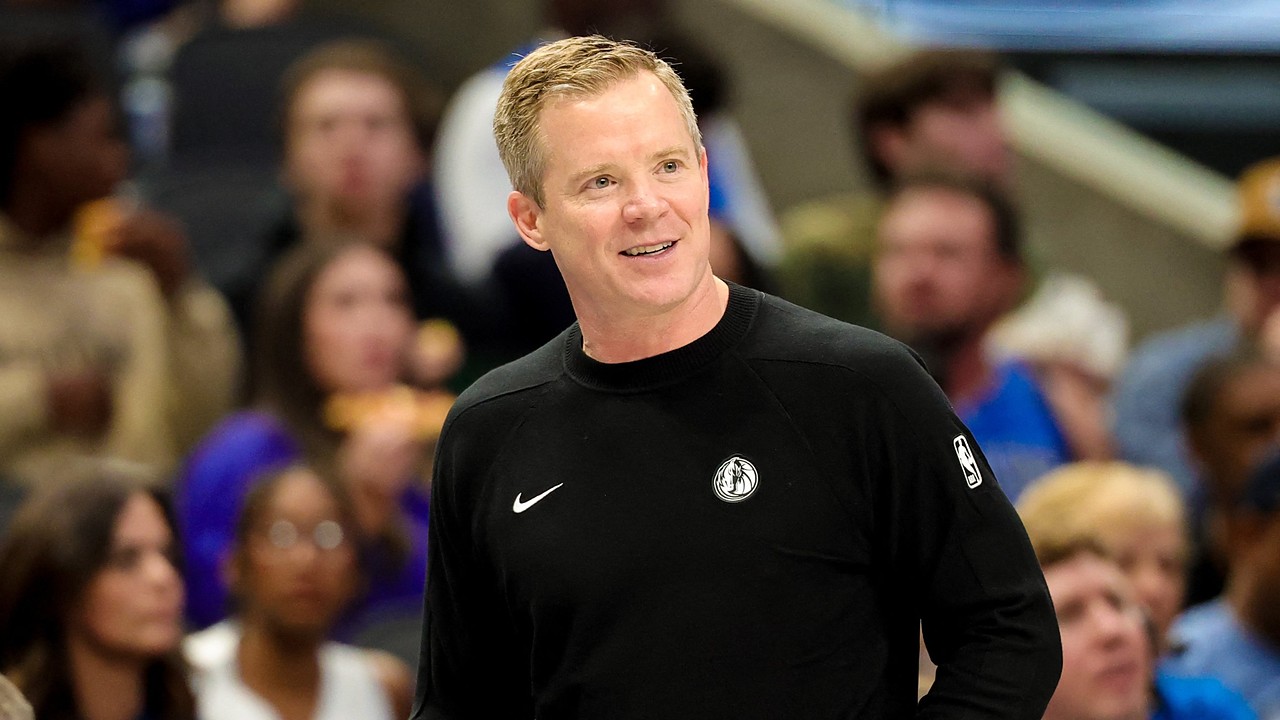 Dallas Mavericks head coach Dusty May smiles on the sideline in a black Mavericks sweater as fans watch behind him.