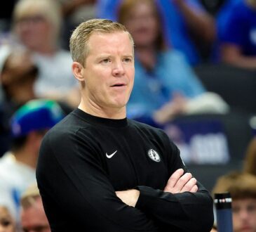 Dallas Mavericks head coach Dusty May stands with his arms crossed on the sideline as fans watch behind him.