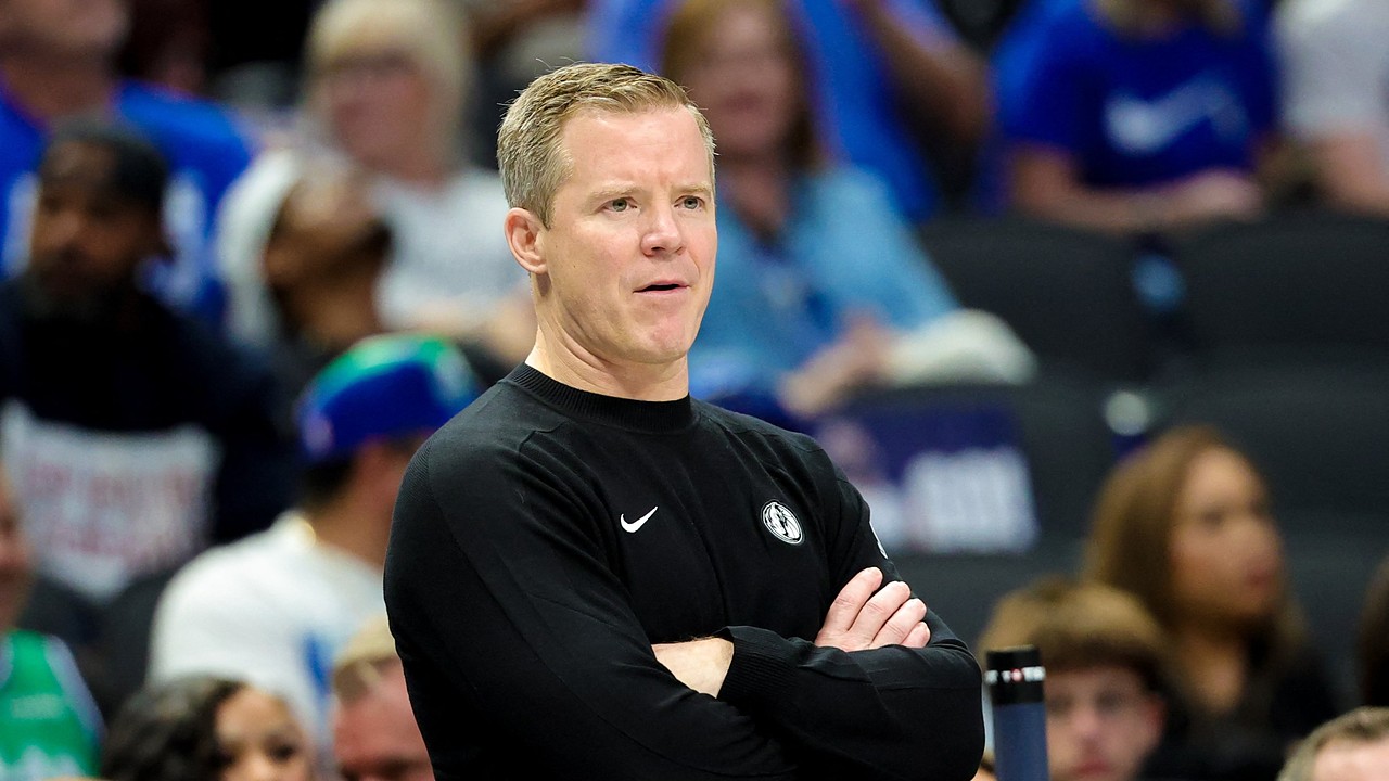 Dallas Mavericks head coach Dusty May stands with his arms crossed on the sideline as fans watch behind him.