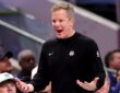 Dallas Mavericks head coach Dusty May reacts on the sideline with his mouth open and both palms turned up as fans look on.
