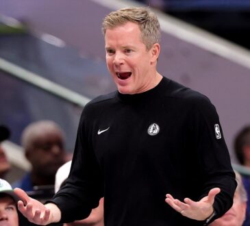 Dallas Mavericks head coach Dusty May reacts on the sideline with his mouth open and both palms turned up as fans look on.
