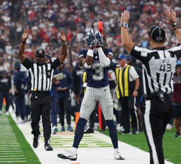 Cowboys wide receiver George Pickens covers his face mask with both hands as two officials raise their arms on the sideline.