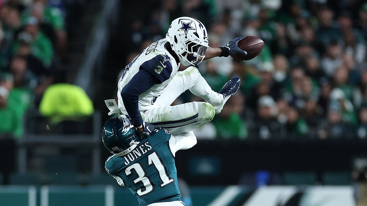 Dallas Cowboys wide receiver George Pickens hurdles over an Eagles cornerback with the ball stretched out in one hand.