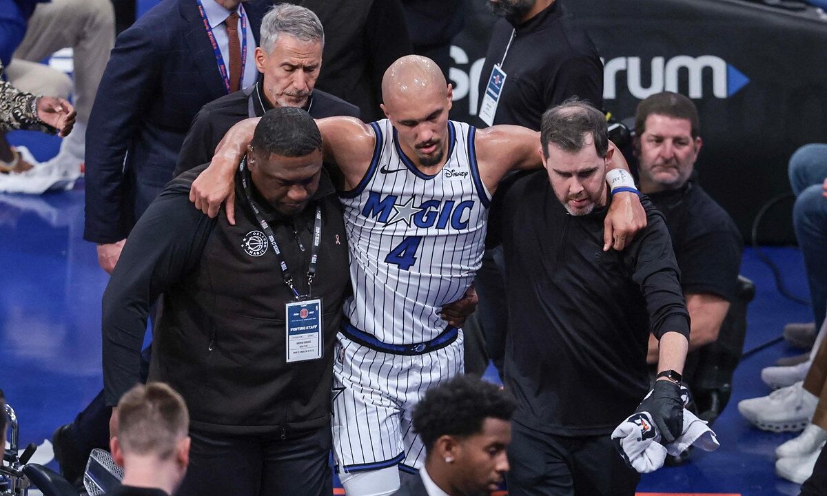 Orlando Magic guard Jalen Suggs is helped off the court by two team staff members, with his arms over their shoulders.
