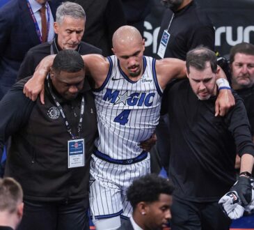 Orlando Magic guard Jalen Suggs is helped off the court by two team staff members, with his arms over their shoulders.