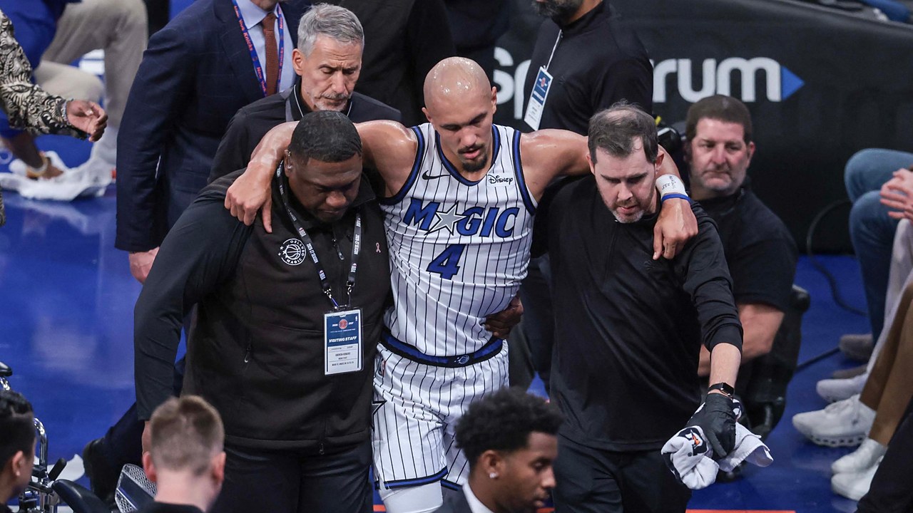 Orlando Magic guard Jalen Suggs is helped off the court by two team staff members, with his arms over their shoulders.
