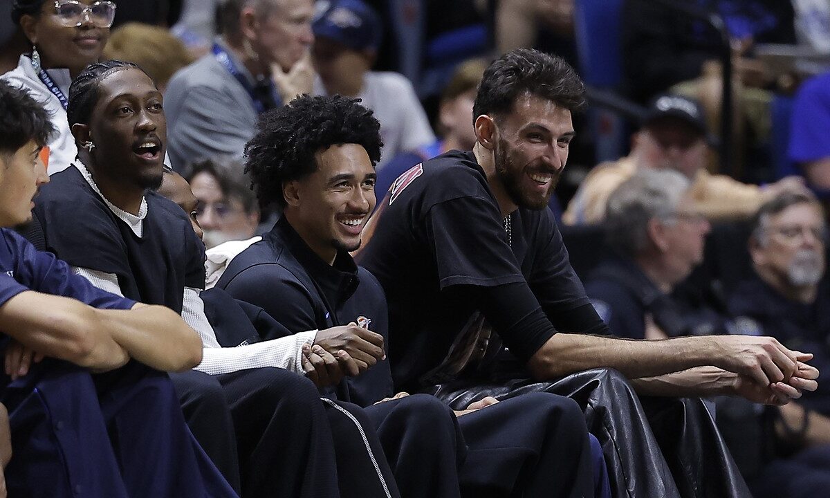 Oklahoma City Thunder players Jalen Williams, Jared McCain, and Chet Holmgren smile while watching from the bench in street clothes.
