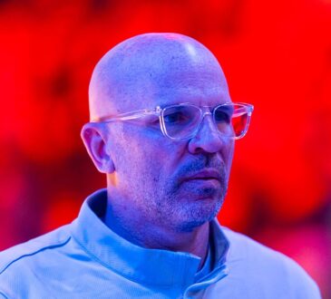 Then-Dallas Mavericks head coach Jason Kidd looks on in clear glasses and a light blue quarter-zip under red arena lights.
