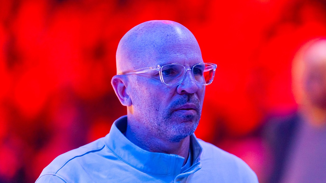 Then-Dallas Mavericks head coach Jason Kidd looks on in clear glasses and a light blue quarter-zip under red arena lights.