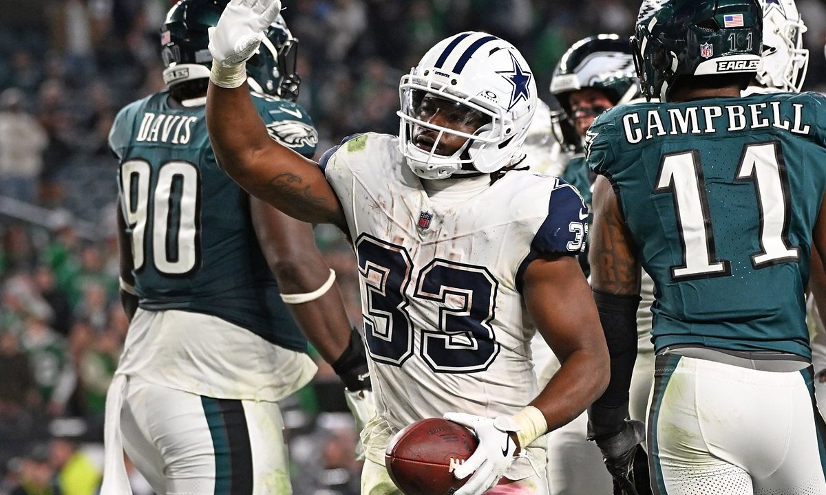 Dallas Cowboys running back Javonte Williams raises his hand and holds the football after scoring, with Philadelphia Eagles players around him.