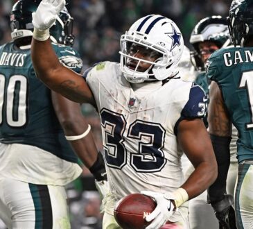 Dallas Cowboys running back Javonte Williams raises his hand and holds the football after scoring, with Philadelphia Eagles players around him.