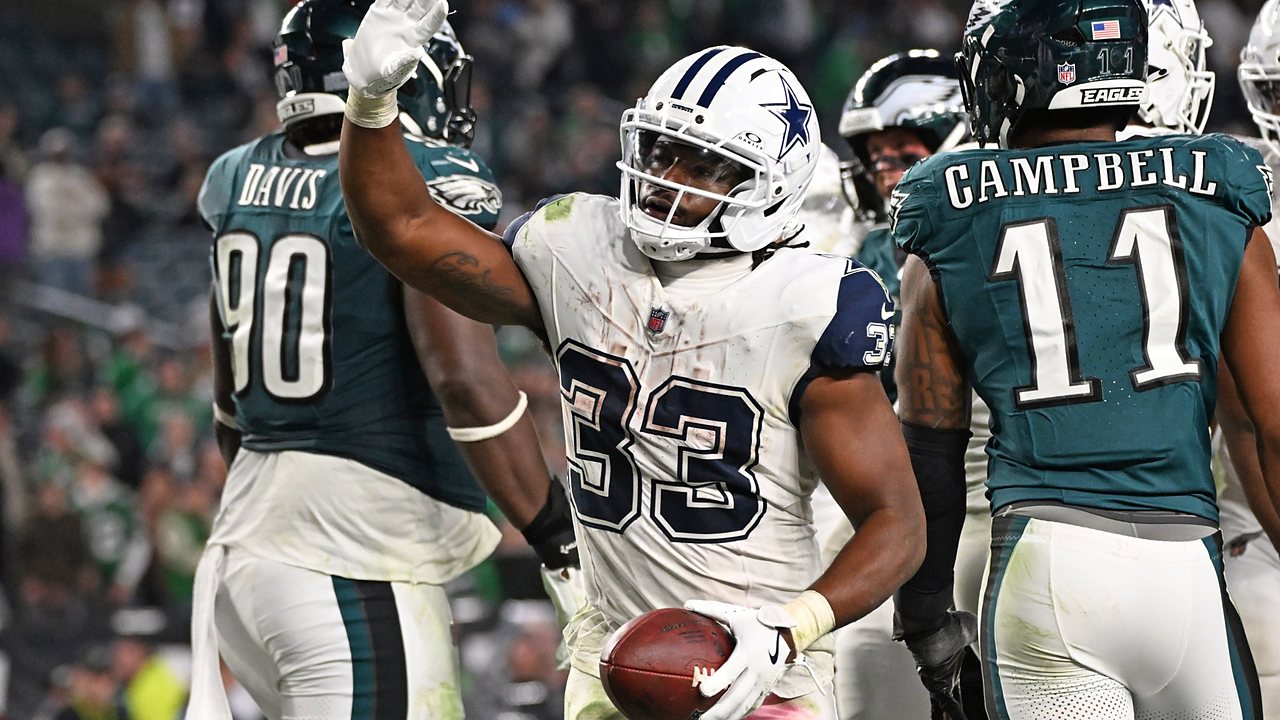Dallas Cowboys running back Javonte Williams raises his hand and holds the football after scoring, with Philadelphia Eagles players around him.