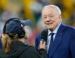 Dallas Cowboys owner Jerry Jones smiles while speaking into a reporter's microphone on the field before a game.