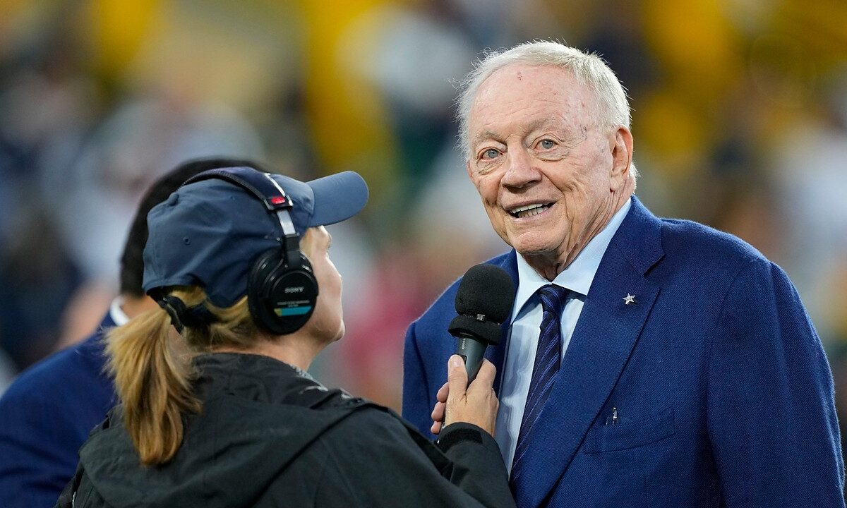 Dallas Cowboys owner Jerry Jones smiles while speaking into a reporter's microphone on the field before a game.
